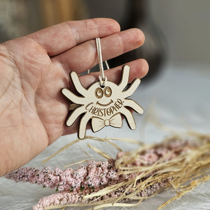 A hand holding a wooden ornament shaped like a smiling spider with the name "Christoph" written on it.