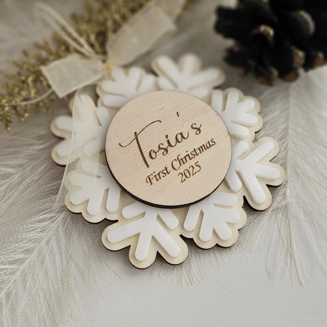 A wooden ornament with the name "Tosia's First Christmas 2015" engraved on it, surrounded by snowflake-shaped ornaments and feathers.