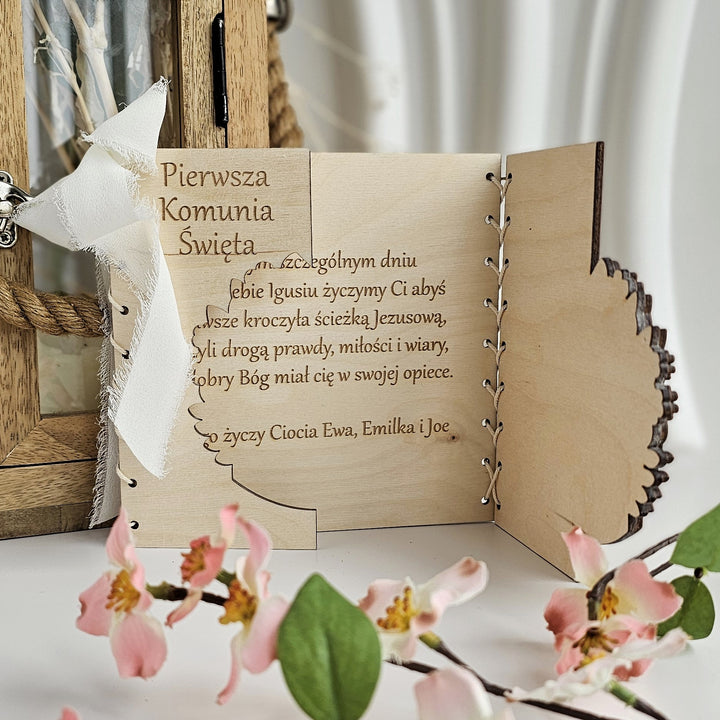 a wooden card with a floral design, placed on a table with pink flowers. The card appears to be a handmade greeting card or a personalized keepsake, with a lace-like texture and a wooden frame.