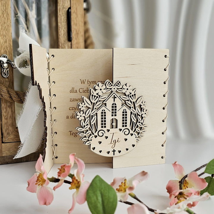 A wooden card with a decorative cutout in the center, featuring a house and flowers, placed on a table with pink flowers in the foreground.