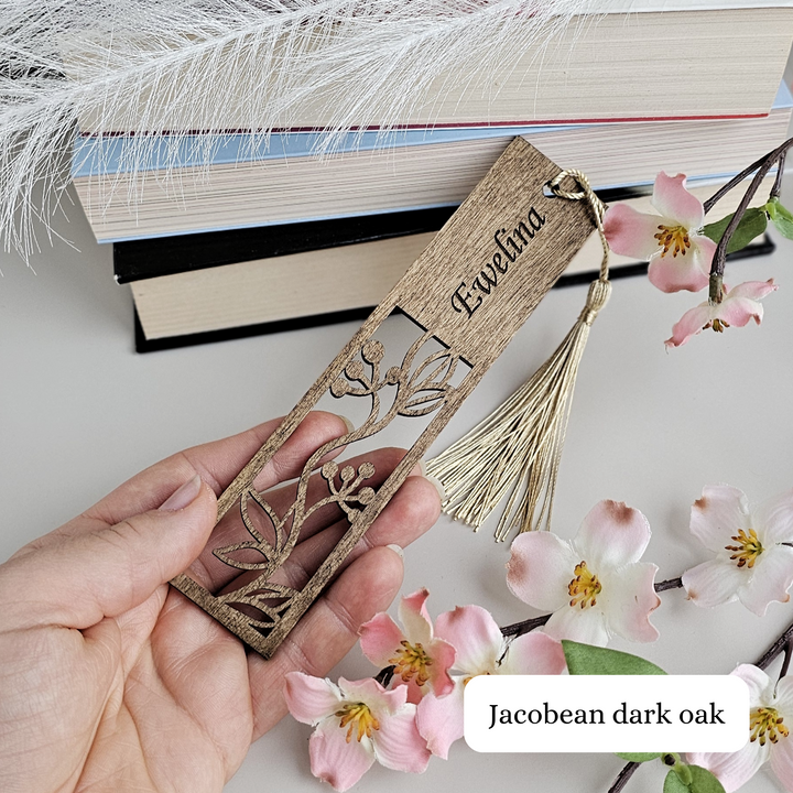 A hand holding a wooden bookmark with a tassel, surrounded by pink flowers and a stack of books.