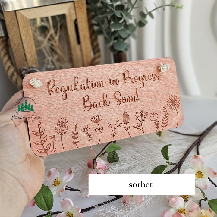 A wooden sign with the text "Regulation in Progress - Back Soon!" is being held in front of a floral arrangement.