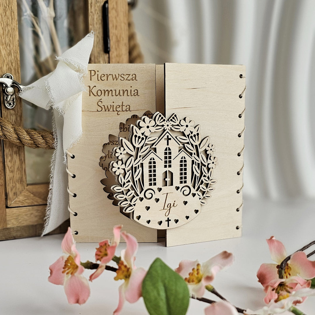 a wooden card with a decorative house design, placed on a table with pink flowers in the foreground.