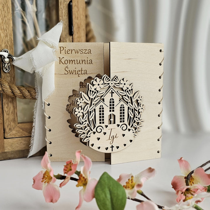 a wooden card with a decorative house design, placed on a table with pink flowers in the foreground.