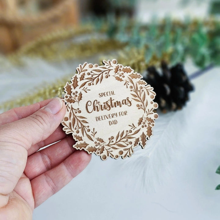 A hand holding a wooden coaster with a Christmas wreath design and text on it.