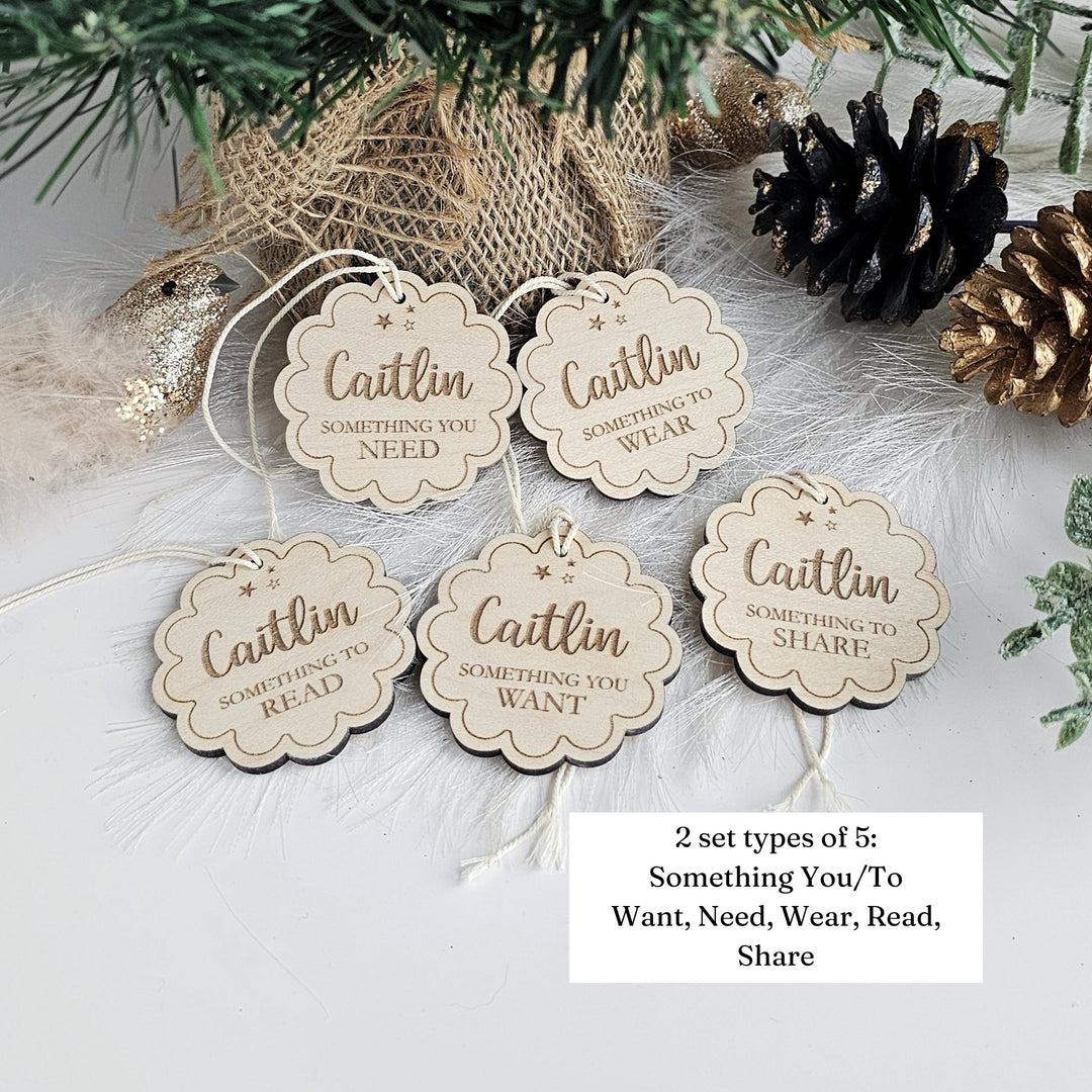 a set of five wooden tags with the text "Caitlin something you need, something you want, something you share" written on them. The tags are placed on a white surface, with pine cones and greenery in the background.