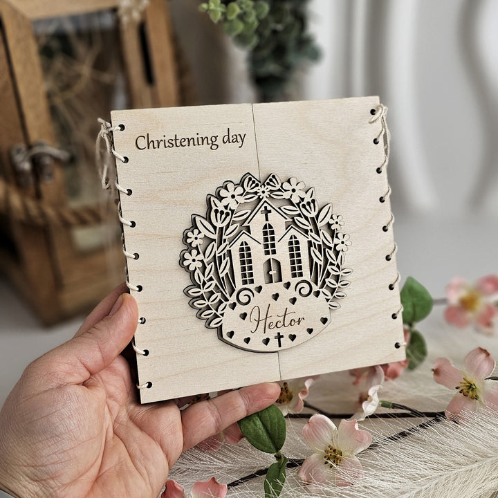 A hand holding a wooden card with a decorative design and the text "Christening day" written on it.