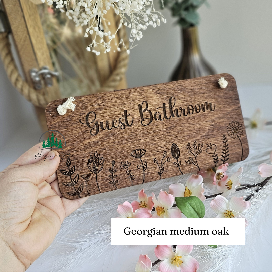 A wooden sign with the text "Guest Bathroom" and decorative floral designs, held in a hand. The sign is surrounded by flowers and a vase, creating a natural and welcoming atmosphere.