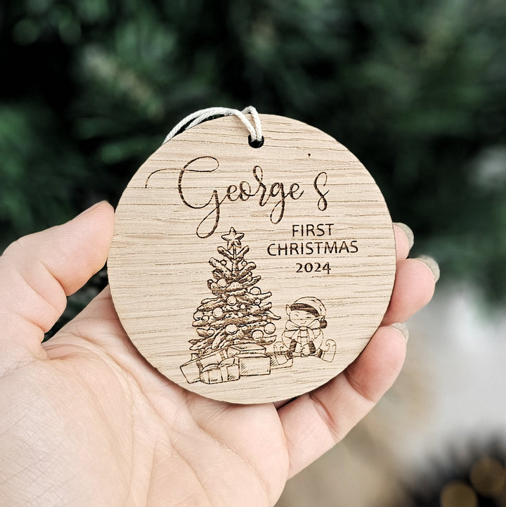 A hand holding a wooden ornament with a Christmas tree and a small figure, along with the text "George's FIRST CHRISTMAS 2023".