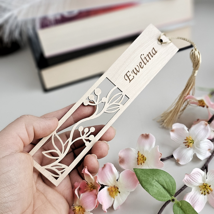 A hand holding a wooden bookmark with a floral design, surrounded by pink flowers and a feather.