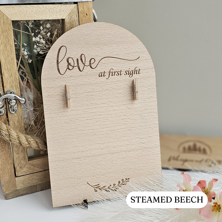 a wooden sign with the text "Love at first sight" engraved on it, along with clothespins. The sign is placed on a table, and there is a small wooden frame and a flower arrangement nearby.