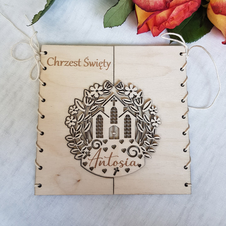 A wooden frame with a decorative design featuring a house and flowers, along with the name 'Antonia' written on it. The frame is placed on a white surface, and there are red and orange flowers in the background.