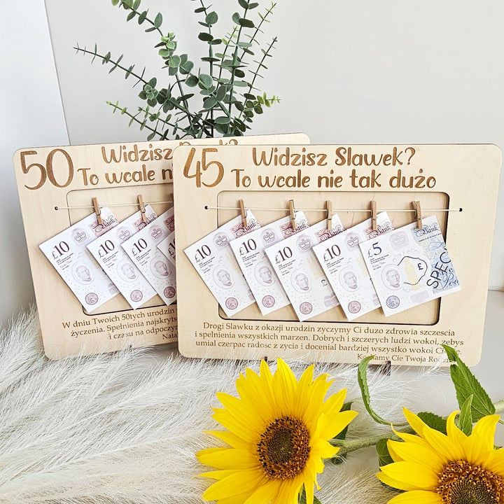 a wooden frame with a plant and sunflowers in the background. The frame has a wooden border and contains several pieces of paper with money, including 50 and 45 Polish zloty notes.