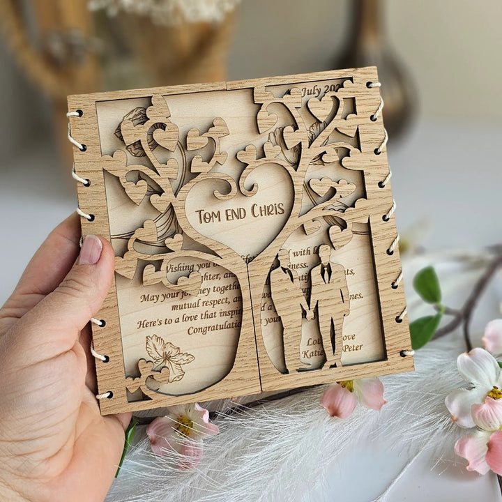 A hand holding a wooden card with intricate designs, including a heart and floral patterns, against a backdrop of flowers and greenery.