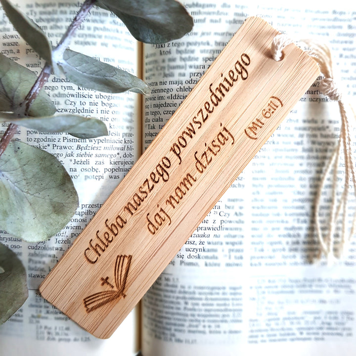 A wooden bookmark with Polish text is resting on an open book, with a branch of greenery visible in the background.