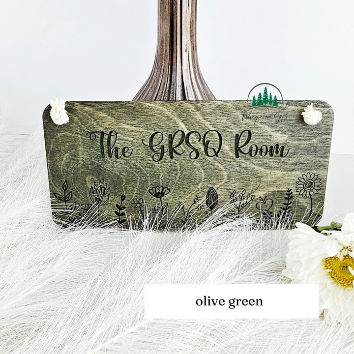 a wooden sign with the text "The GRSQ Room" and decorative elements such as flowers and leaves.