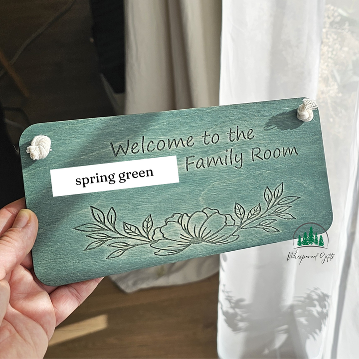 A wooden sign with the text "Welcome to the Family Room" and a floral design, held in someone's hand.