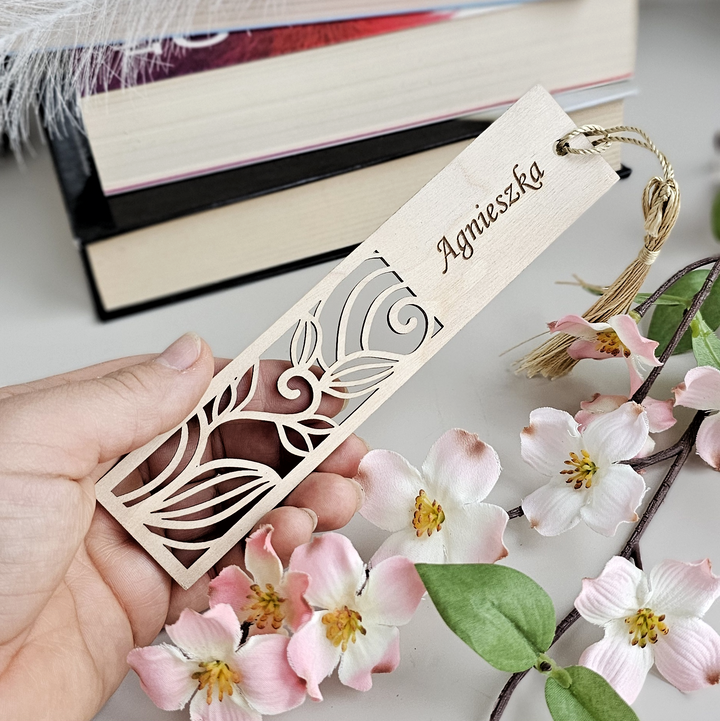 A hand holding a wooden bookmark with a floral design, surrounded by pink flowers and a feather.