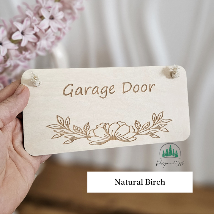 A wooden sign with the text "Garage Door" and a floral design, held in a hand against a background of pink flowers.