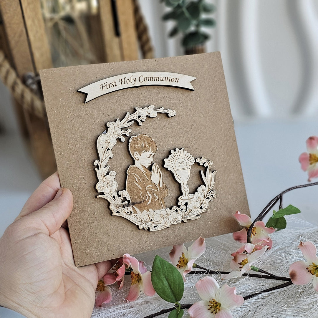 A hand holding a card with a wooden cutout of a child praying, surrounded by a wreath of flowers and pink blossoms.