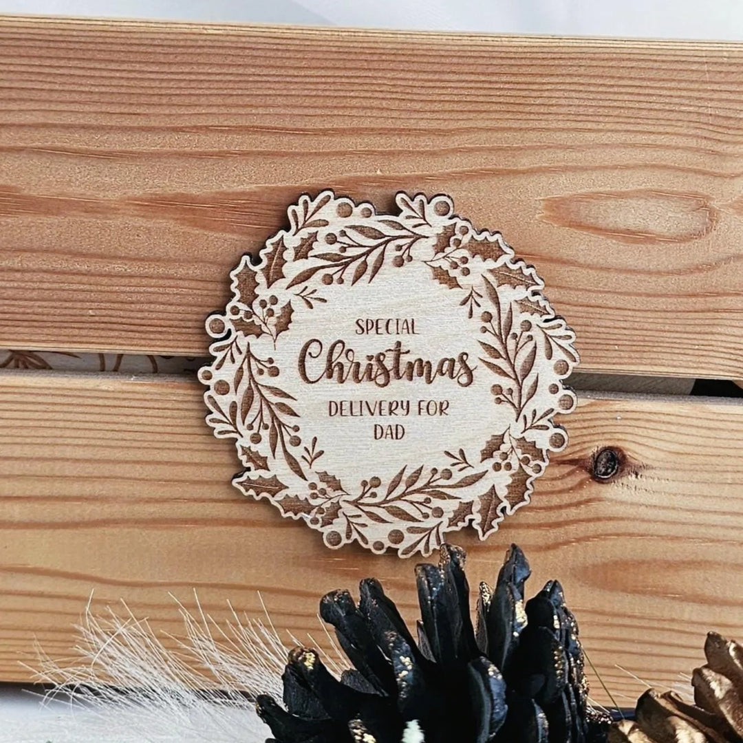 A wooden wreath-shaped coaster with the text "Special Christmas Delivery for Dad" engraved on it, placed on a wooden surface.