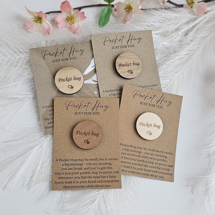a set of four wooden "Pocket Hug" magnets, each with a unique message engraved on it. The magnets are displayed on a white background, with a floral arrangement in the background.