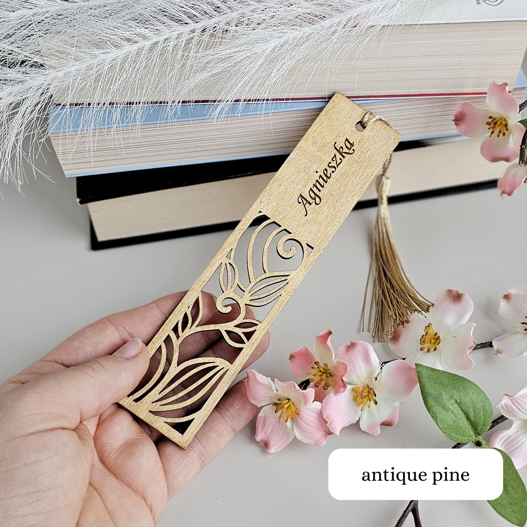 A hand holding a wooden bookmark with a floral design, surrounded by books and pink flowers.