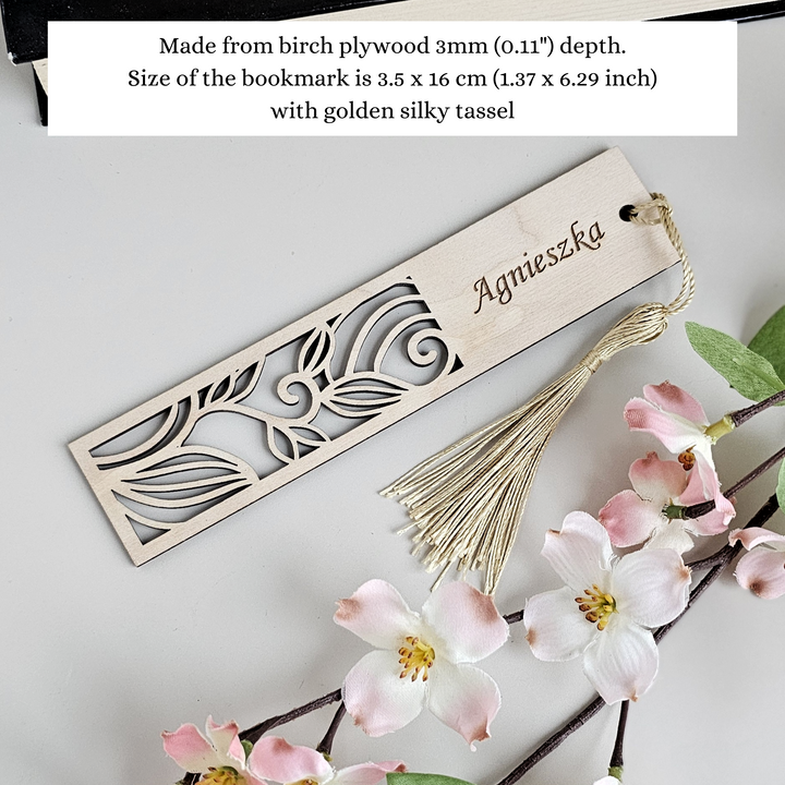 A wooden bookmark with a decorative cutout design and a tassel, placed next to a branch with pink flowers.