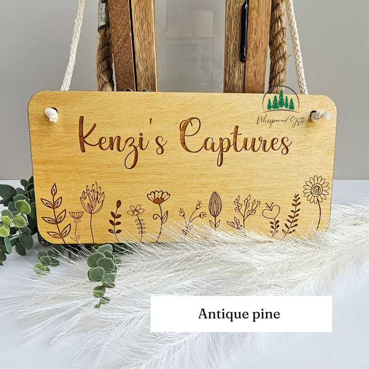a wooden sign with the name "Kenzi's Captures" engraved on it, surrounded by various floral and plant illustrations. The sign is placed on a white surface, with some greenery visible in the background.