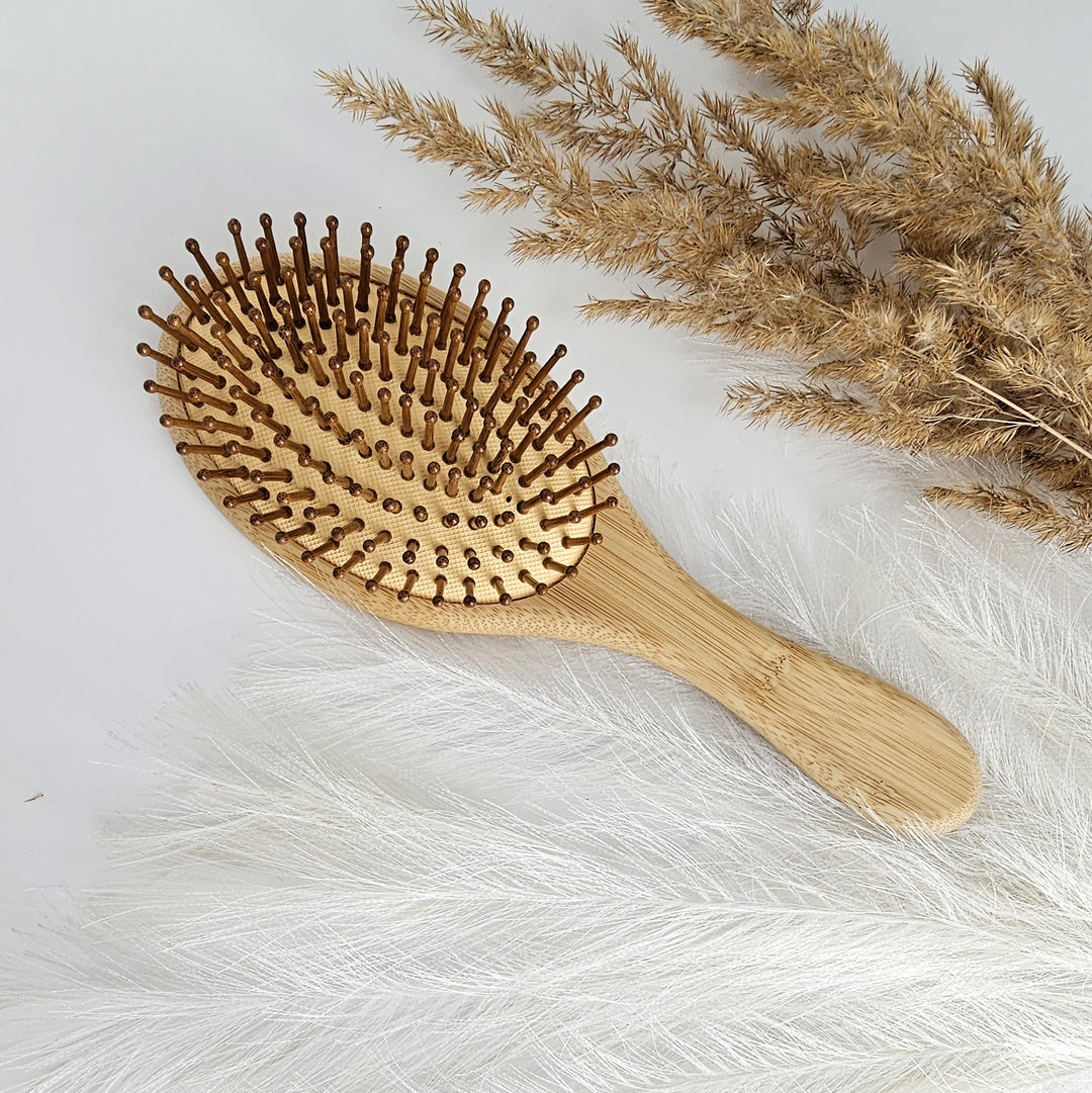 A wooden hairbrush with a natural wooden handle and bristles is placed on a white surface, accompanied by dried plant material.