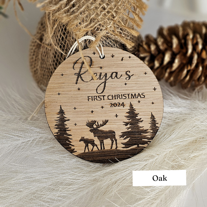 A wooden ornament with a deer silhouette and a starry night sky, featuring the text "Reya's FIRST CHRISTMAS 2024 Oak" and a pine cone in the background.