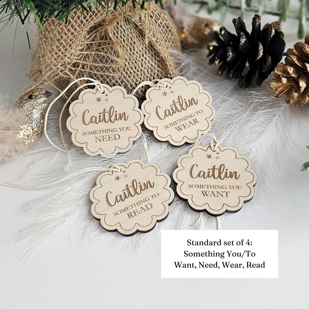 A set of four wooden tags with the text "Caitlin Something You Need Something You Want Something You Wear, Read" on them, placed on a white surface next to a burlap sack and pine cones.