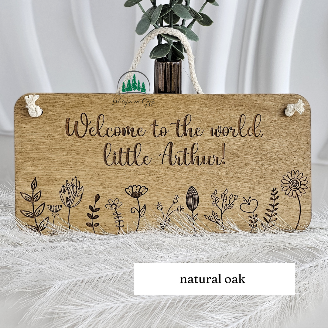 A wooden sign with the text "Welcome to the world, little Arthur!" written on it, along with various floral and plant illustrations. The sign is placed on a white surface, and there is a vase with greenery and a rope nearby.