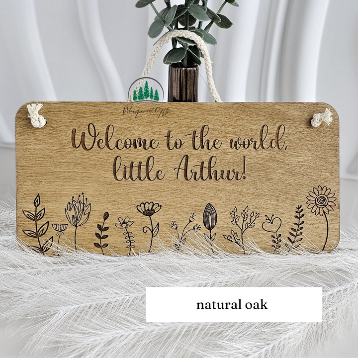 A wooden sign with the text "Welcome to the world, little Arthur!" written on it, along with various floral and plant illustrations. The sign is placed on a white surface, and there is a vase with greenery and a rope nearby.