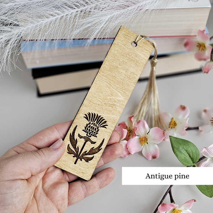 A hand holding a wooden bookmark with a decorative design, surrounded by books and pink flowers.