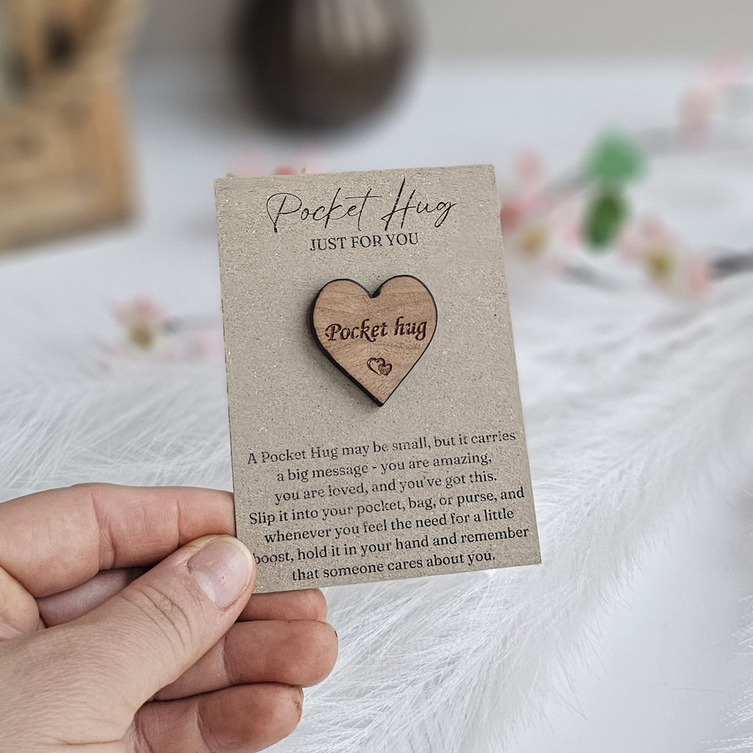 A hand holding a wooden heart-shaped object with a message about a "Pocket Hug" on a card.