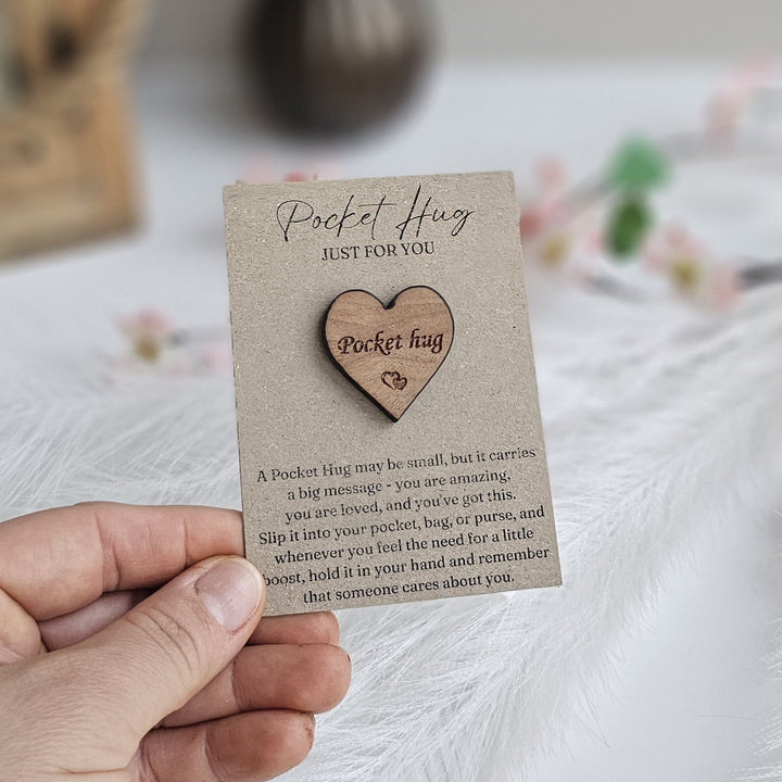 A hand holding a wooden heart-shaped object with a message about a "Pocket Hug" on a card.