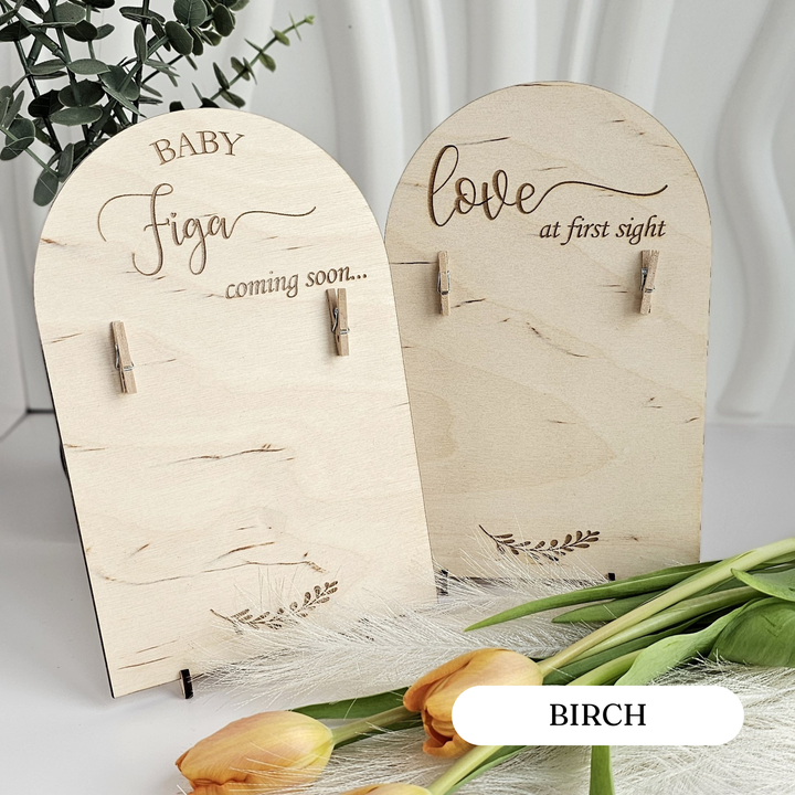 two wooden signs with the words "Baby Figa" and "Love at first sight" written on them, along with clothespins. The signs are placed on a white surface, and there are some flowers and greenery in the background.