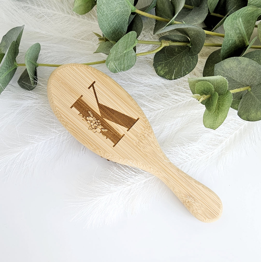 A wooden brush with a letter "K" engraved on it, placed on a white surface next to some green leaves.