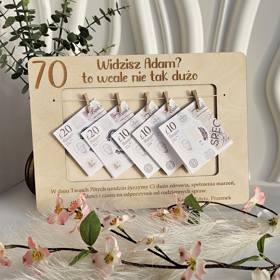 A wooden frame with a series of Polish banknotes hanging from it, placed on a white surface with a plant and flowers in the background.