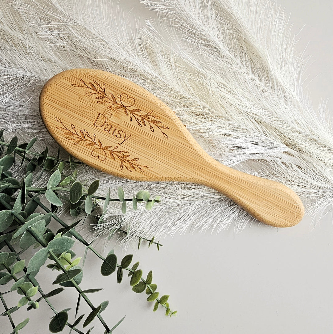 A wooden brush with the name "Daisy" engraved on it, placed on a white surface next to some greenery.