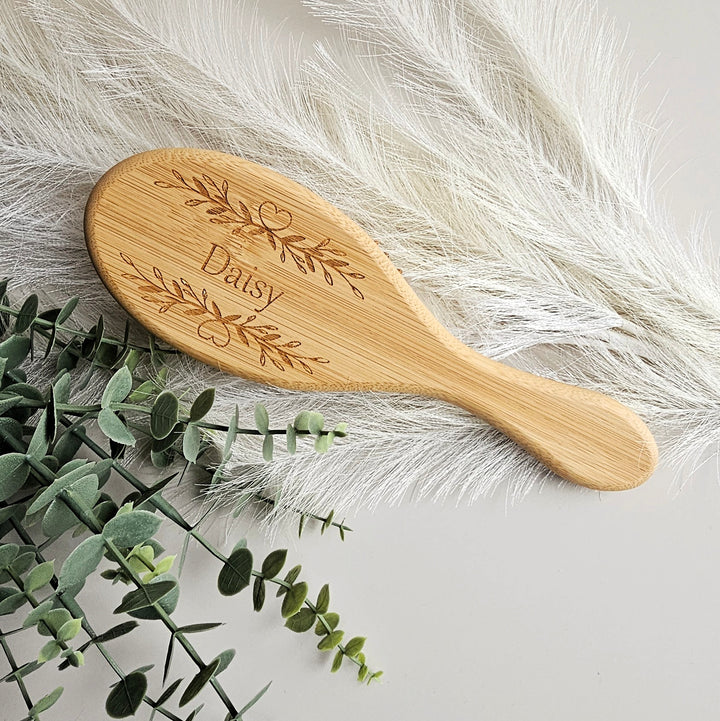 A wooden brush with the name "Daisy" engraved on it, placed on a white surface next to some greenery.
