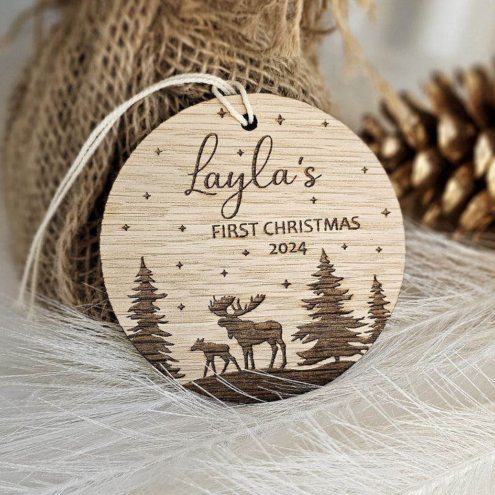 A wooden ornament with a deer silhouette and pine trees, featuring the text "Layla's FIRST CHRISTMAS 2023" engraved on it.