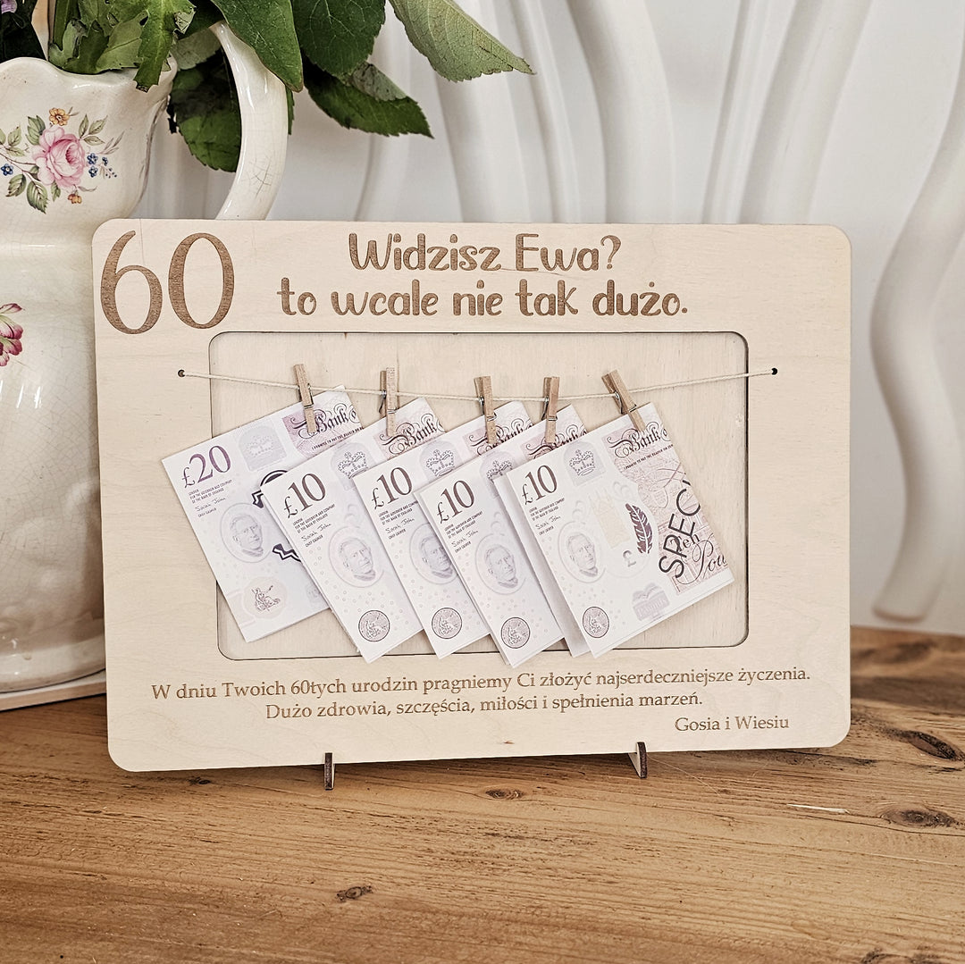A wooden frame with money notes hanging from it, placed on a wooden surface next to a white vase with flowers.