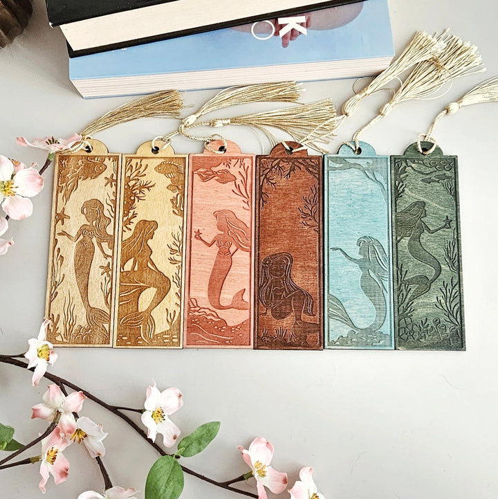 A collection of six mermaid-themed bookmarks, each with a unique design, arranged on a white surface. The bookmarks are made of wood and feature illustrations of mermaids in various colors and poses.