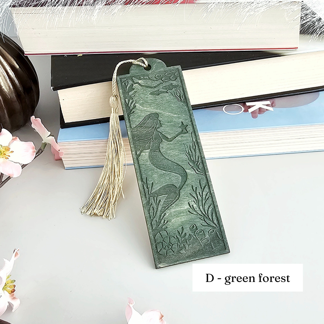 A green bookmark with a mermaid design is placed on top of a stack of books, surrounded by pink flowers.