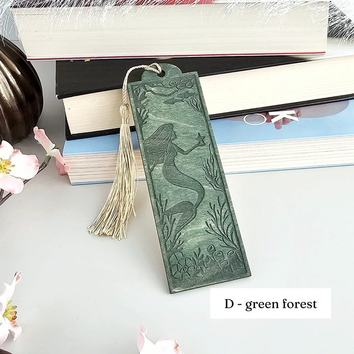 A green bookmark with a mermaid design is placed on top of a stack of books, surrounded by pink flowers.