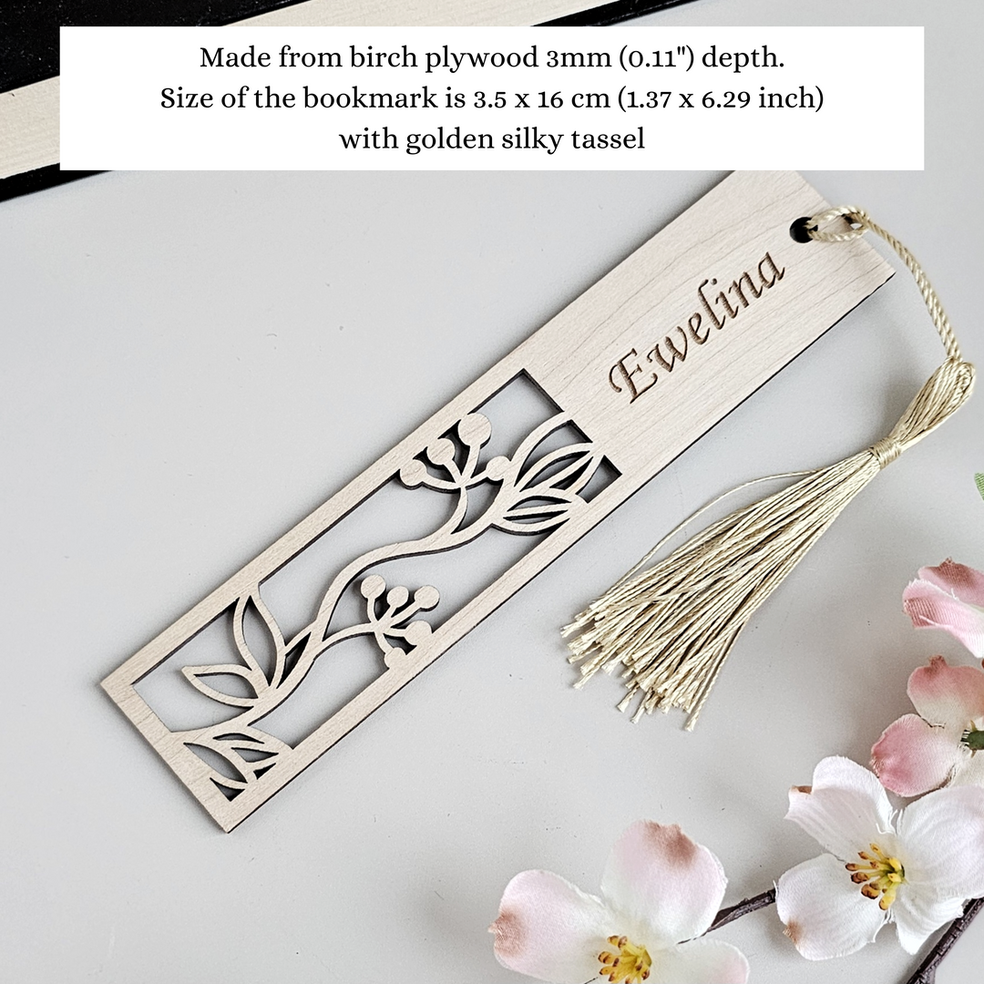 A wooden bookmark with a tassel and a small floral design, placed on a white surface next to pink flowers.