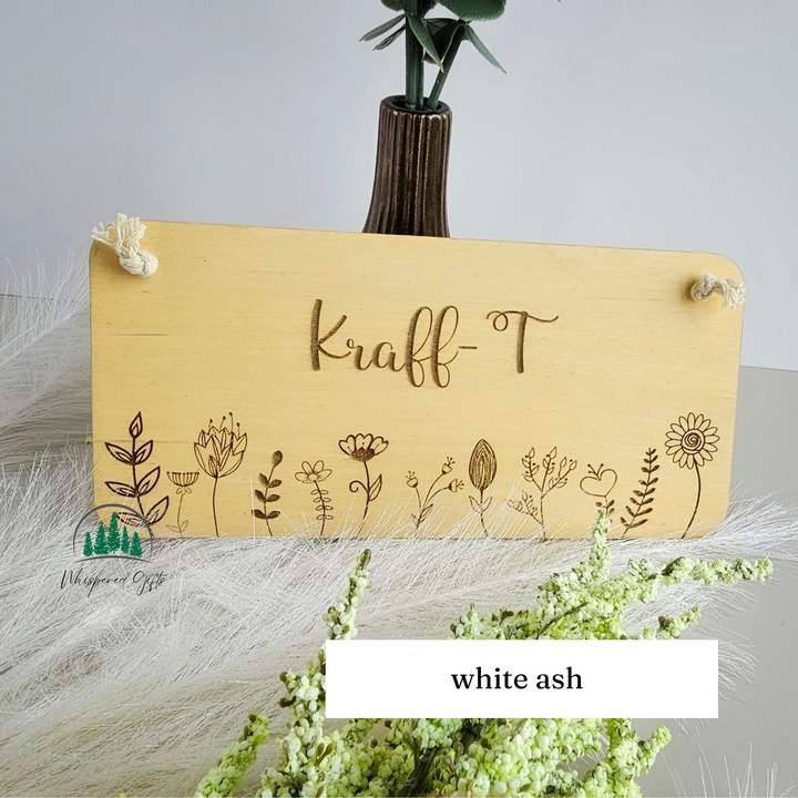 a wooden sign with the name "Kraff-T" engraved on it, surrounded by various floral designs. The sign is placed on a white surface, with a vase of greenery and white flowers in the background.