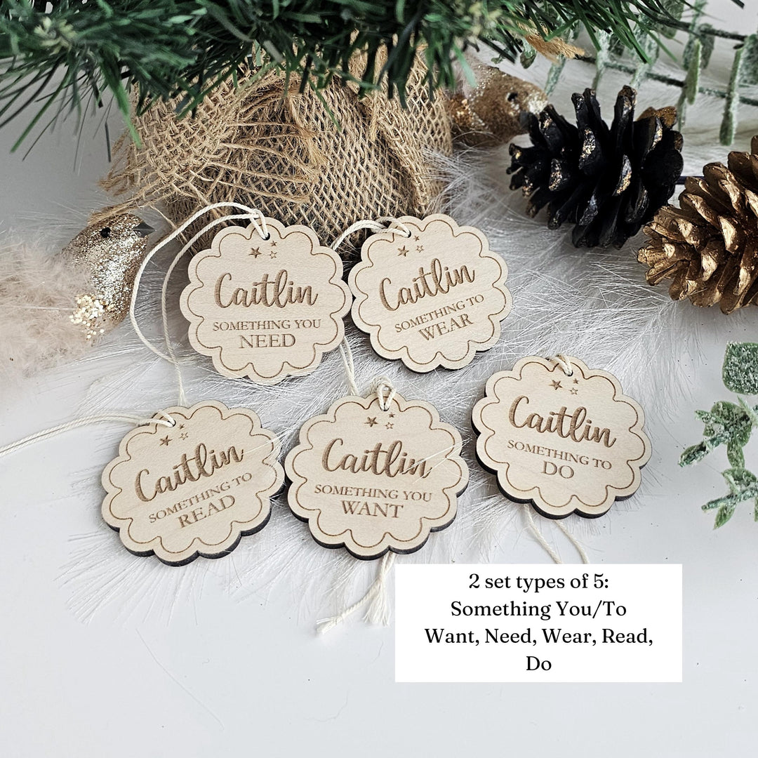 a set of five wooden tags with the text "Caitlin something you need, something you want, something you read, something you wear, something you do" written on them. The tags are arranged on a white surface, with a pine tree branch and pine cones in the background.